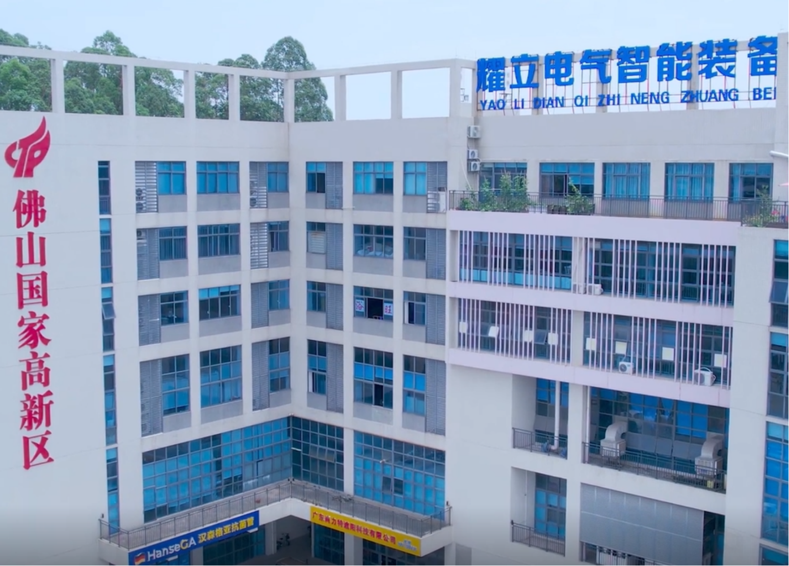 现代化厂房外景，灰色建筑配蓝色玻璃幕墙，周围绿树环绕，阳光充足